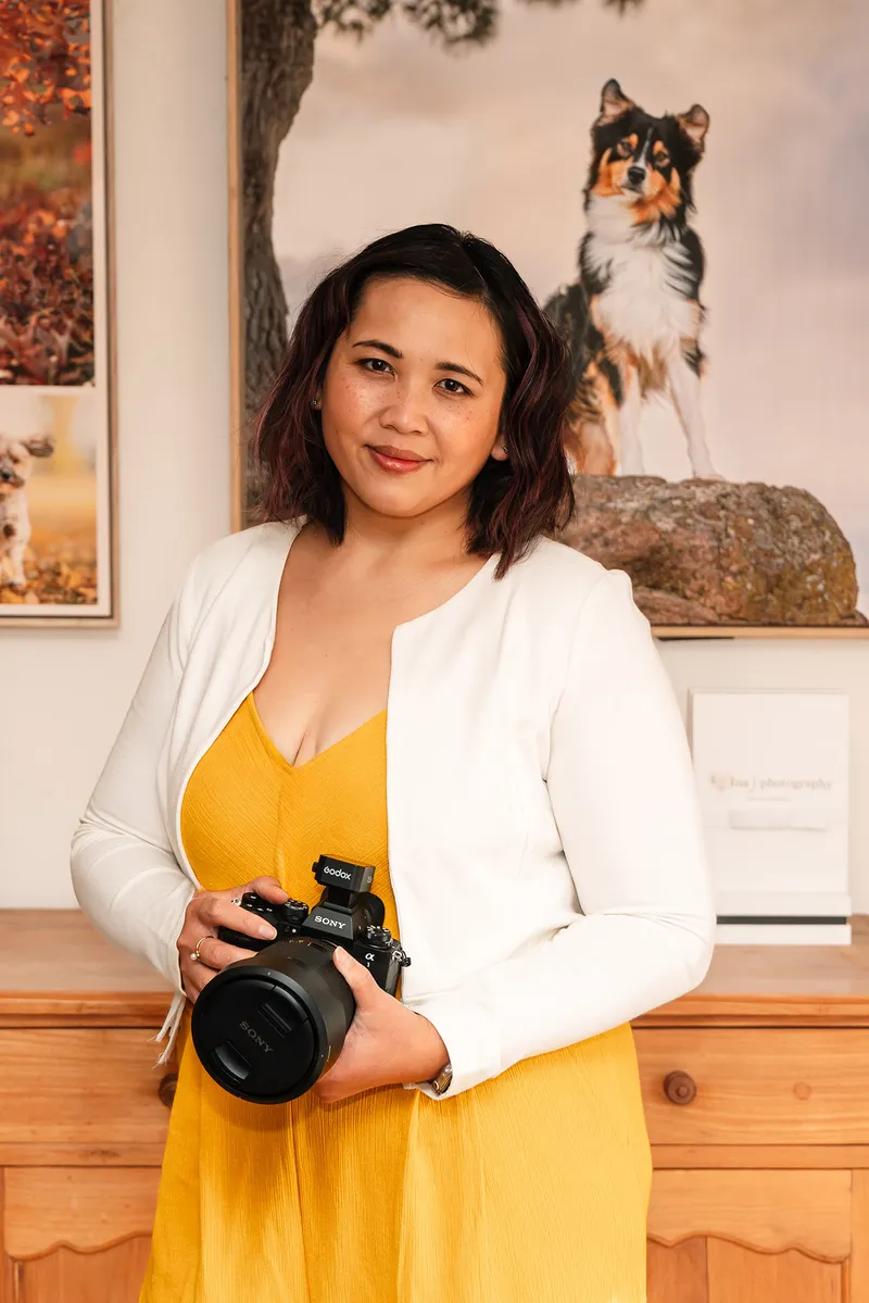 Pet photography business coach Ina J in a yellow dress holding her camera in her studio, framed dog prints on the wall behind her