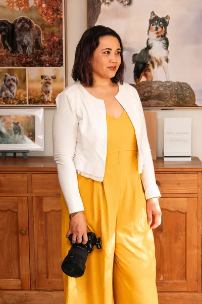 Pet photography business coach Ina J holding her camera in her studio, framed prints on the wall behind her